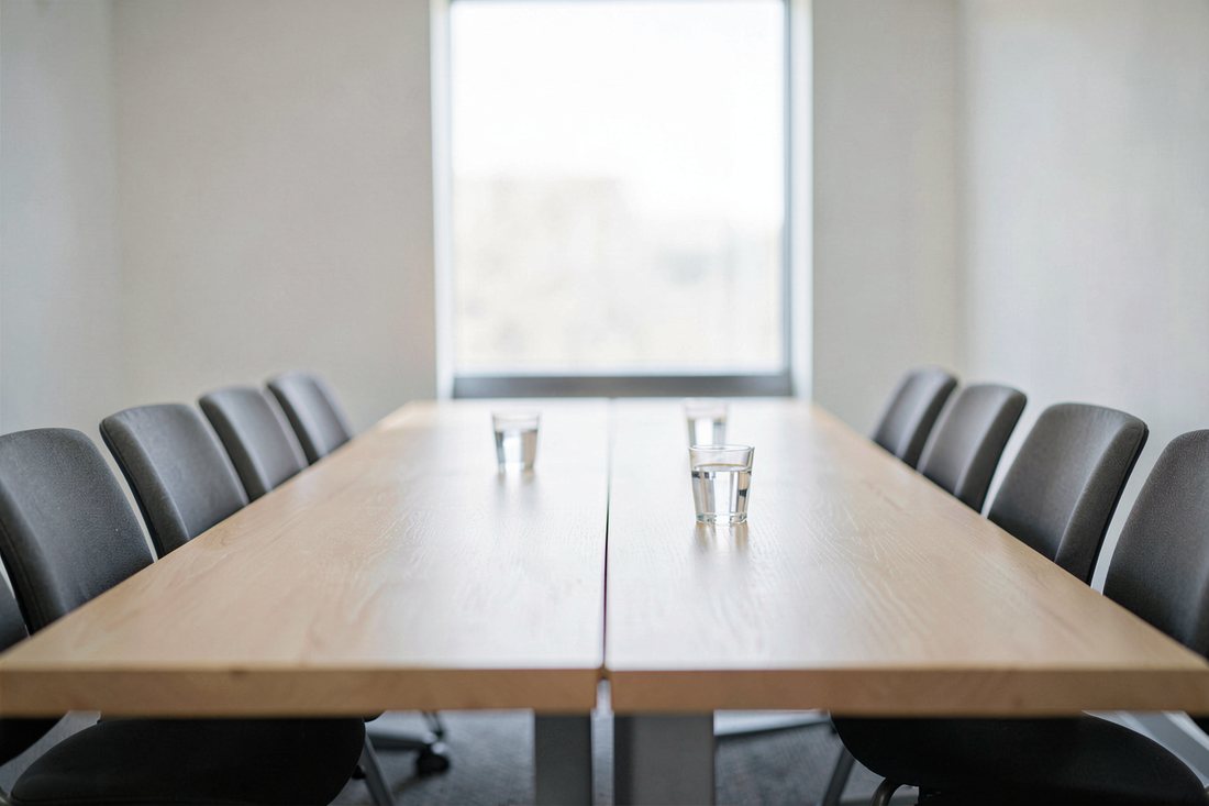 An empty bright meeting room with two light oak tables joined edge to edge along a visible seam, rows of empty grey chairs on both sides, two glasses of water on the table and a tall window